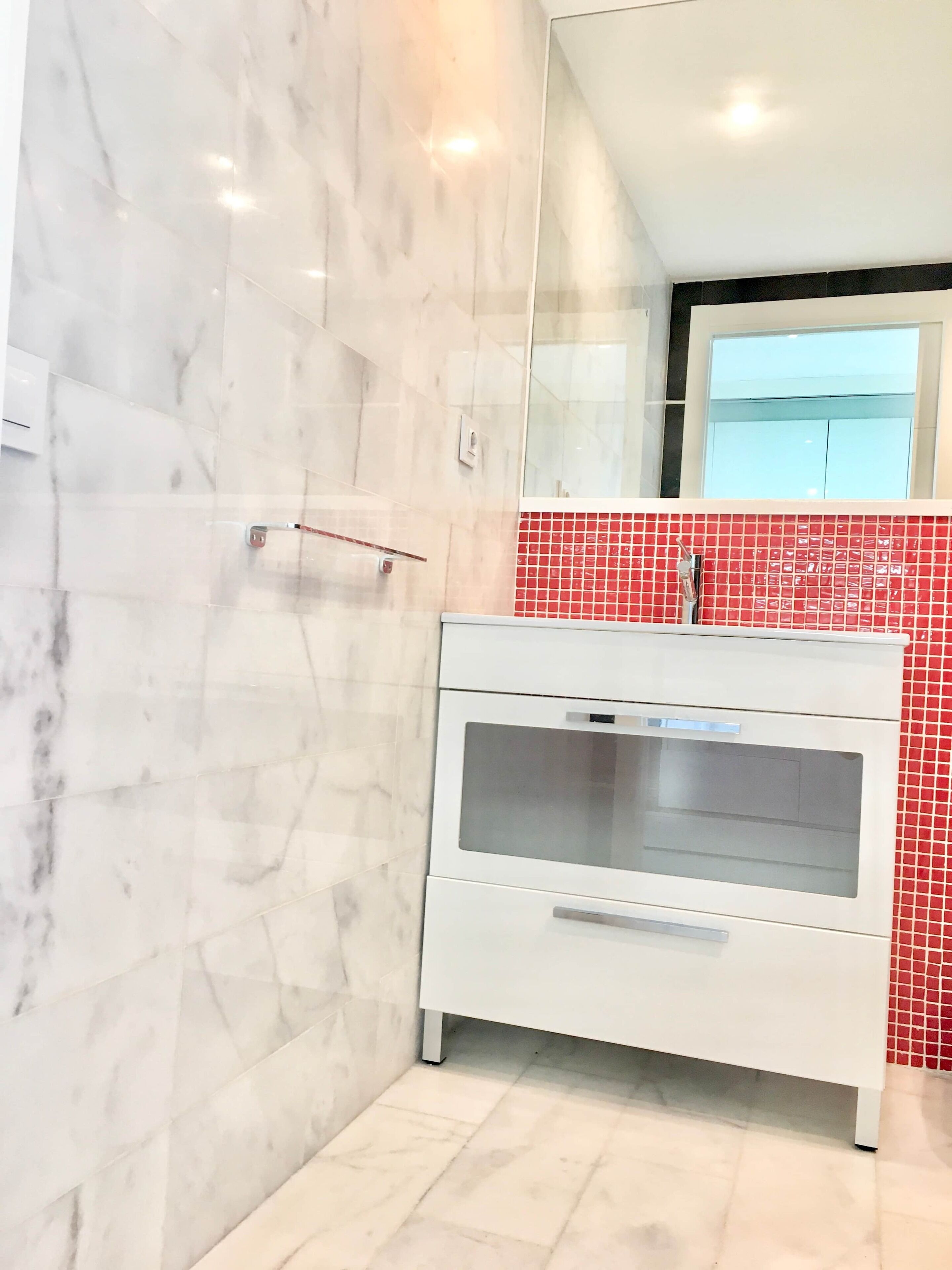 Een badkamer met marmeren wanden, een wit wastafelmeubel, een backsplash met rode tegels en een grote spiegel.