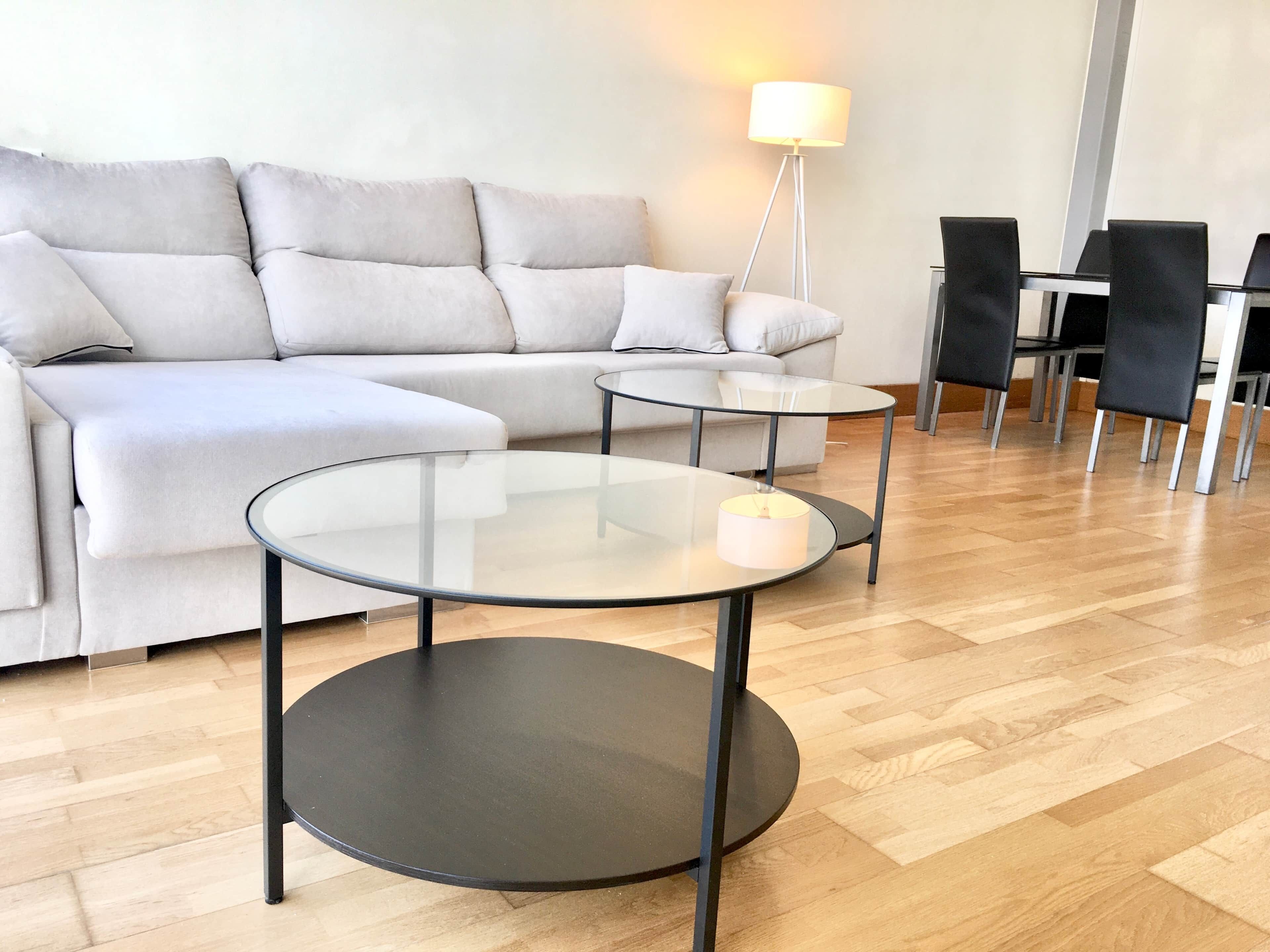 Un salon moderne comprenant un canapé d’angle, deux tables avec plateaux en verre, un lampadaire et un coin repas avec des chaises.