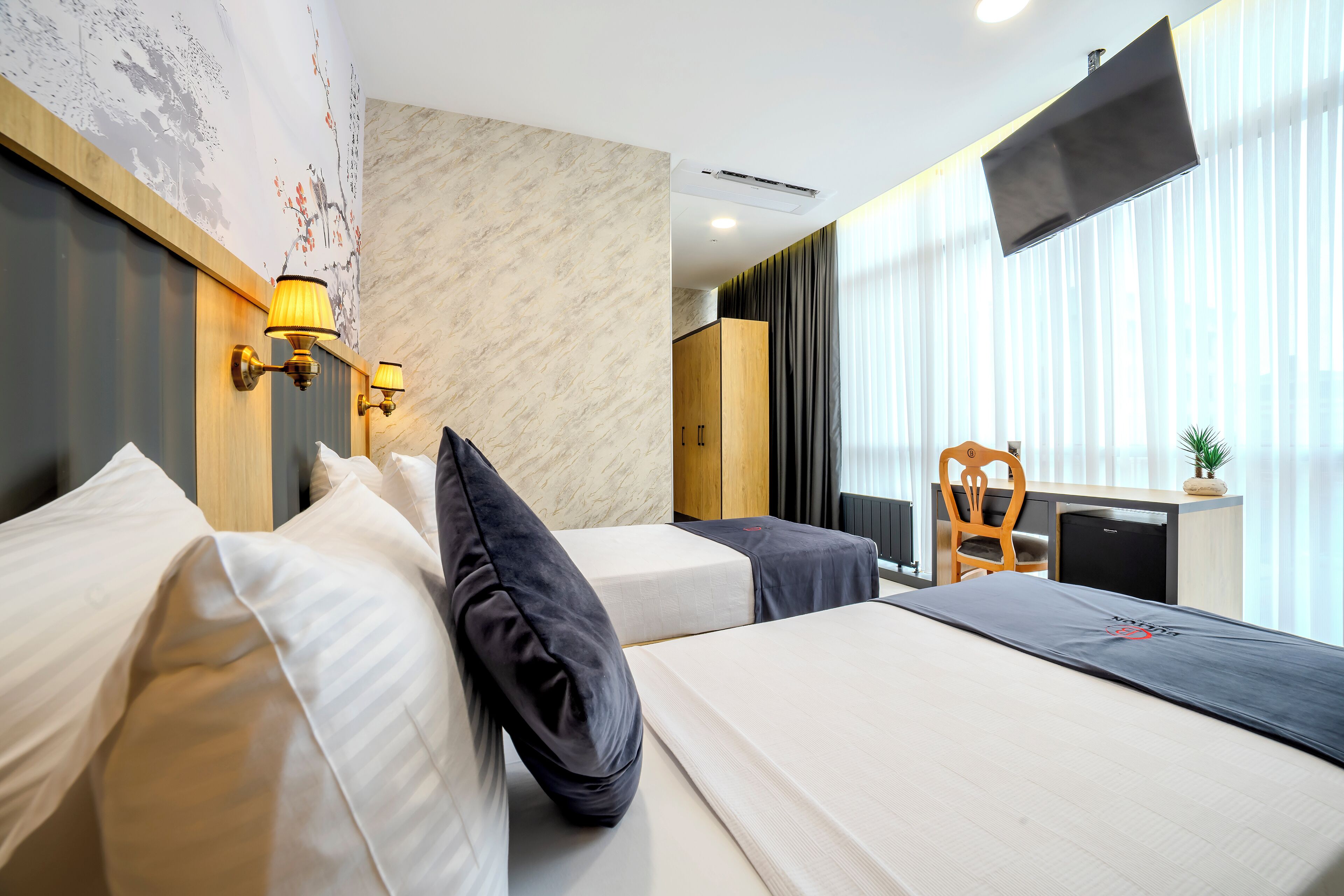 A hotel room with two beds, a desk, a chair, a TV, and a wall with a mural.