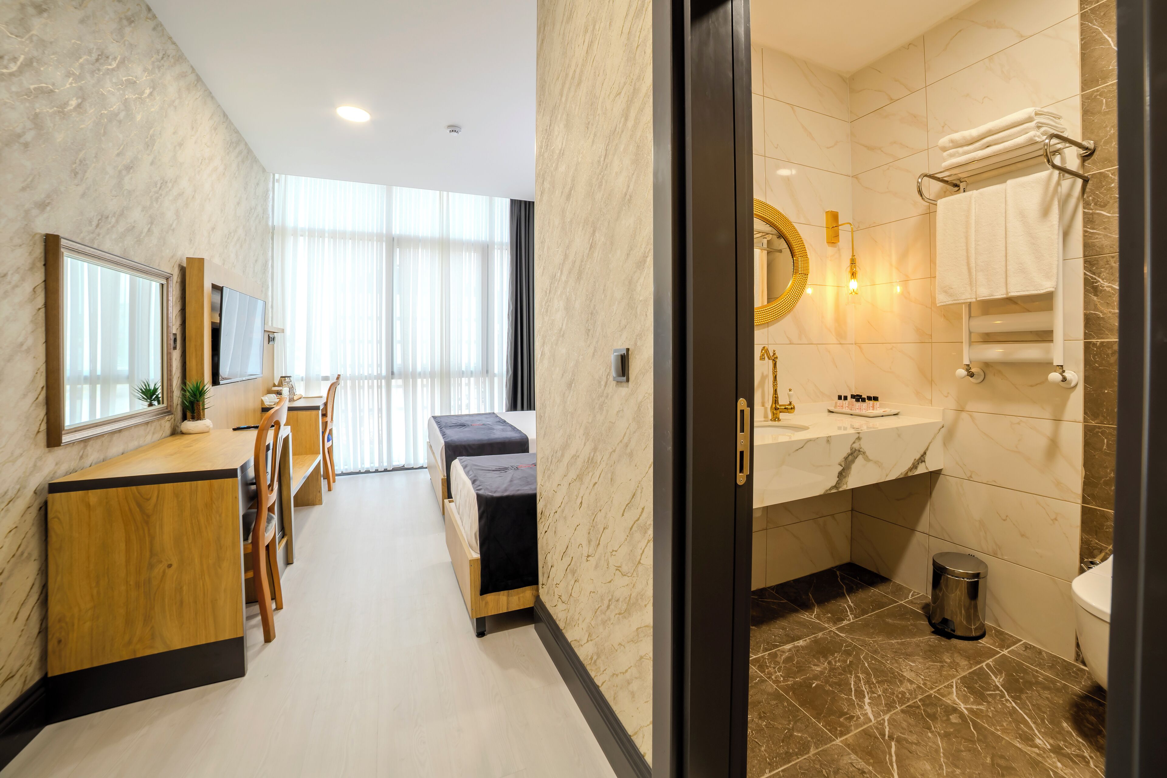 A hotel room with a wooden desk, two beds, a large mirror, and a bathroom with a marble sink and toilet.