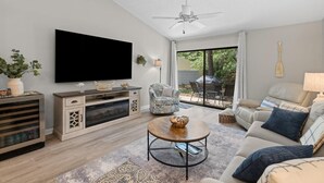 Una sala moderna con un televisor de pantalla plana, un ventilador de techo, un sofá, una mesa de centro y una zona de comedor con asientos exteriores.