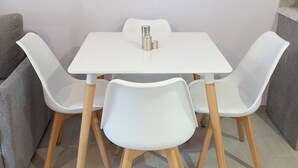 Una mesa blanca con cuatro sillas blancas de patas de madera, un objeto metálico pequeño sobre la mesa y un sofá gris al fondo.