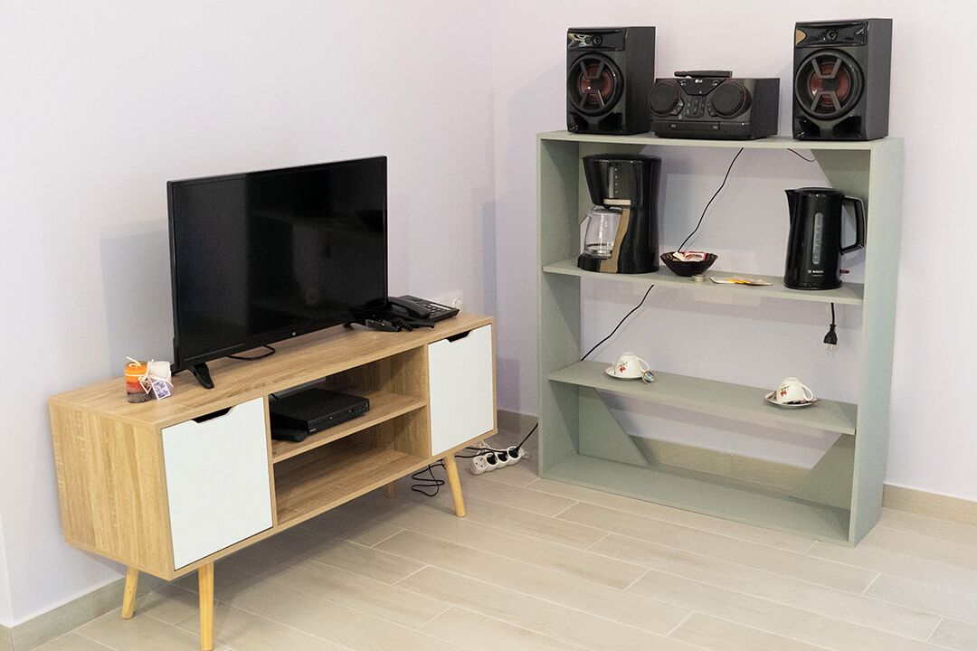 A modern living room with a flat-screen TV on a wooden TV stand, a coffee maker, and a kettle on a green shelf unit.