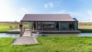 Une maison moderne dotée d’une grande terrasse, avec un bateau amarré à proximité, et un champ verdoyant en arrière-plan.