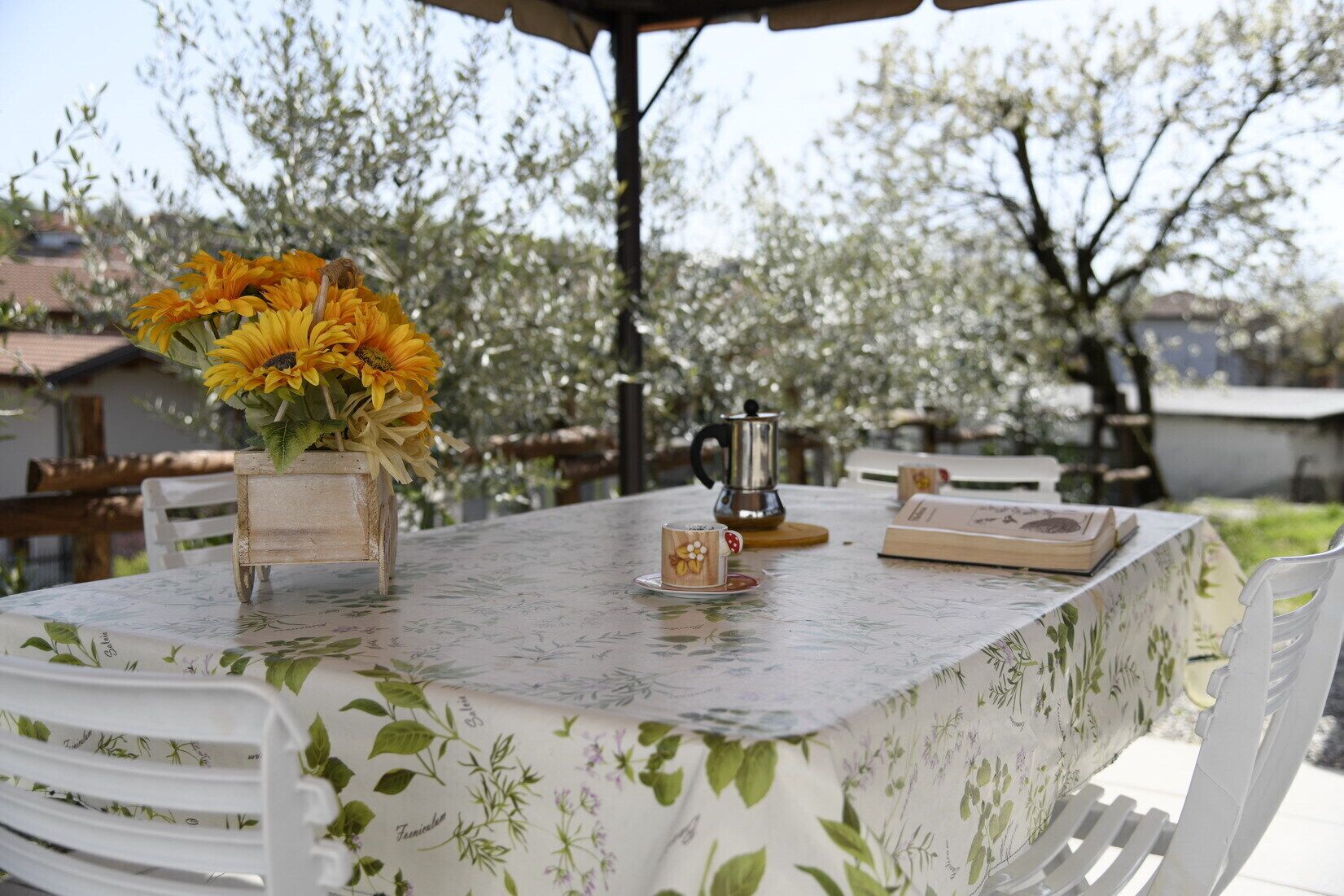An outdoor dining area with a table set for two, a coffee pot, and a vase with sunflowers.