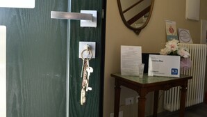 Zona de recepción de un hotel con una puerta verde, una llave colgada en la puerta, una mesa de madera con un cartel de bienvenida para 'Cascina Riva' y un espejo en la pared.