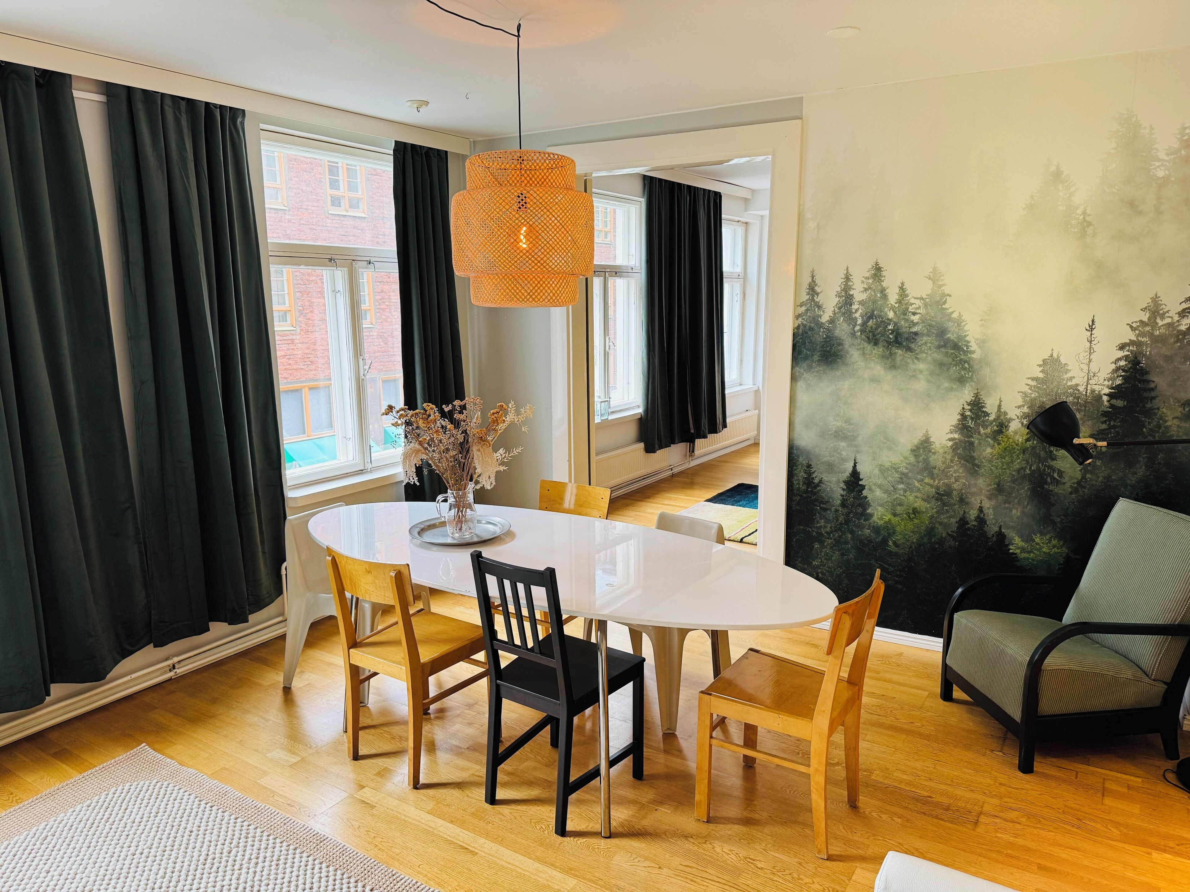 Een eethoek met een witte tafel, zwarte en houten stoelen, een vaas met gedroogde bloemen en een bosdecoratie op de muur.