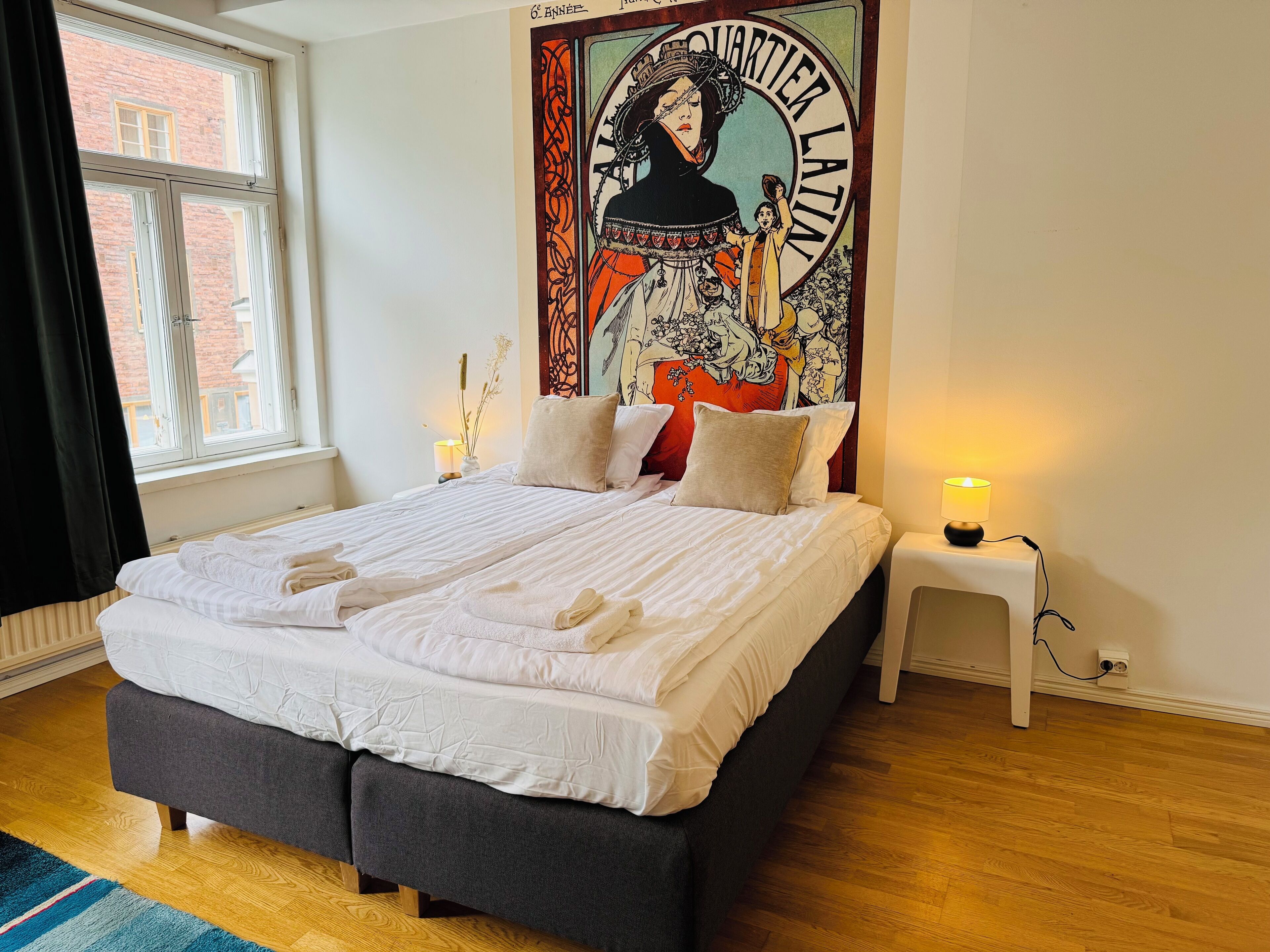 A neatly made bed with white linens, a dark grey upholstered headboard, and a small bedside table with a lamp. A large, colorful tapestry hangs on the wall behind the bed.