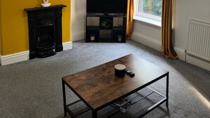 Une pièce avec un mur jaune, un meuble de rangement noir pour appareils électroniques, une cheminée, une table basse en bois et une fenêtre avec des rideaux.