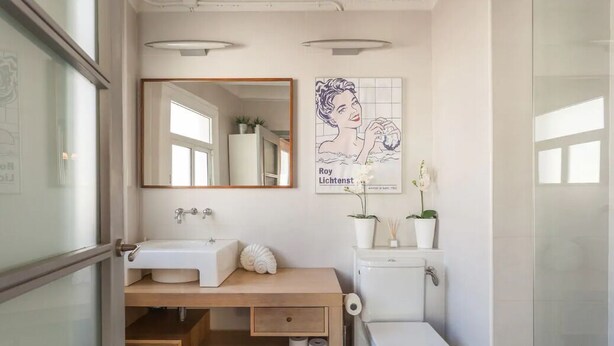 Een moderne badkamer met een houten wastafelmeubel, een witte wastafel, toilet en een grote spiegel. Op de muur hangt een poster en op het meubel staat een pot met een plant.