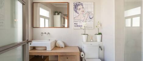 A modern bathroom with a wooden vanity, a white sink, a toilet, and a large mirror. There is a poster on the wall and a potted plant on the vanity.