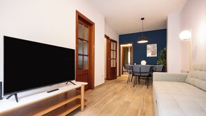 A modern living room with a flat-screen TV on a wooden stand, a grey sofa, a dining table with chairs, and a blue accent wall.