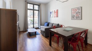 A modern living room with a sofa, a dining table, and red chairs. There is a window with curtains, a ceiling light, and a wall with a menu poster.