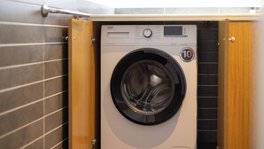 A front-loading washing machine with a control panel, a 10-year warranty badge, and a wooden cabinet door.