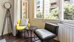 A room with a yellow armchair, a black leather bench, a tripod, and a window with a view of a building and greenery.