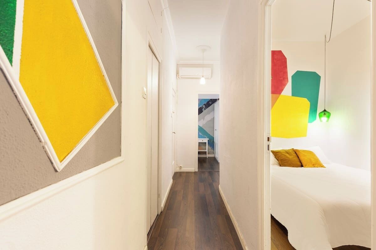 Un couloir étroit au plancher de bois mène à une pièce où se trouve un lit et des œuvres d’art murales aux motifs géométriques et aux couleurs vives.