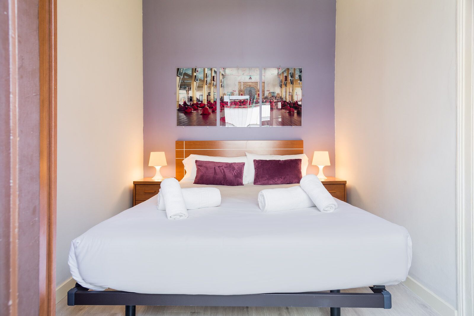 A hotel room with a large bed, two bedside lamps, a headboard with a mirror, and two artwork pieces above the bed.