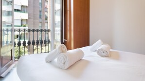 Habitación de hotel con una cama grande, dos toallas enrolladas, una ventana con vistas a edificios y un balcón con barandilla de metal.