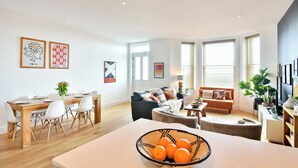 A modern living room with a dining area, featuring a wooden dining table, white chairs, and a large window with blinds. - Stunning 2 Bed Apartment on the beach by Urban Roost (Worthing)