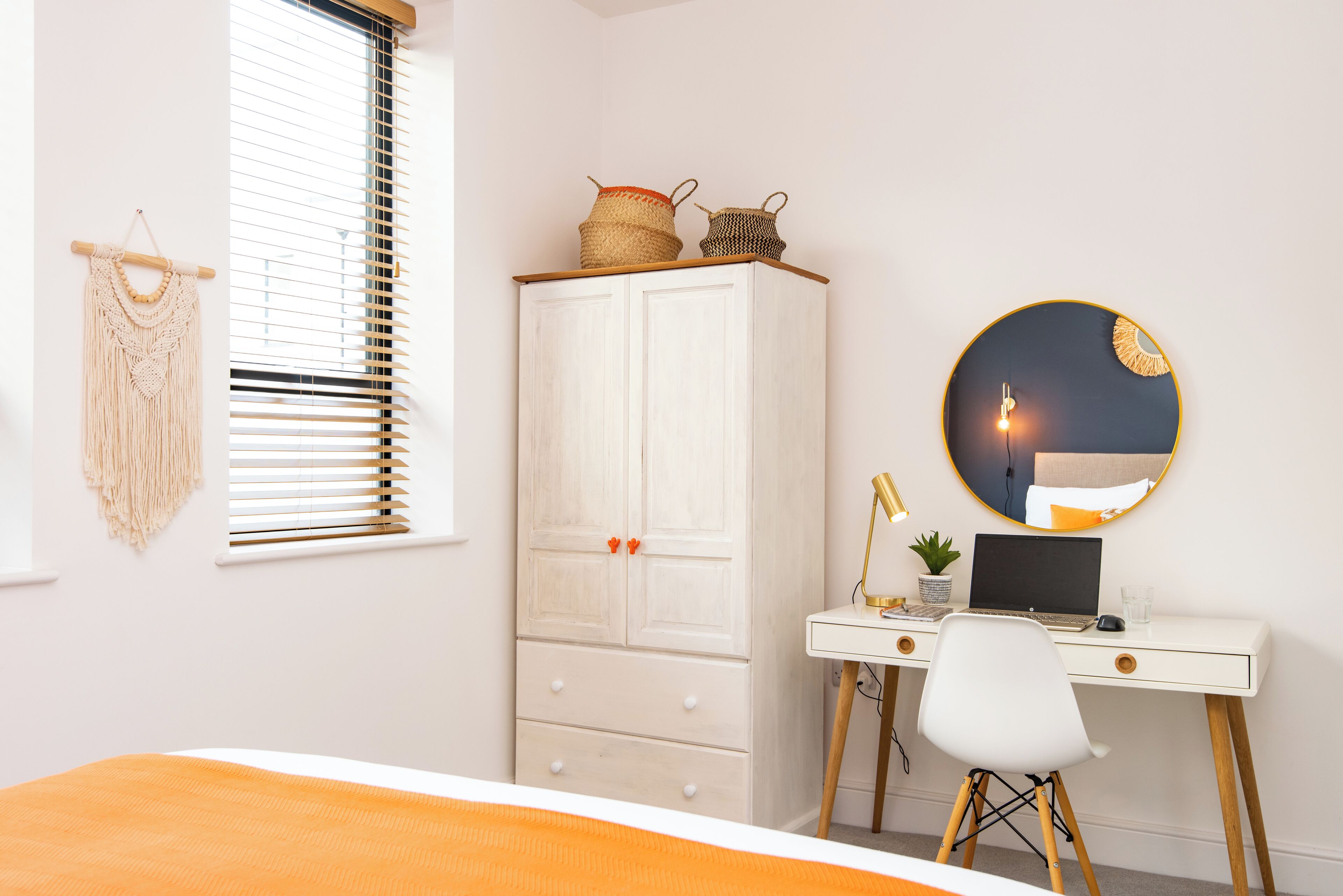 A bedroom with a bed, a desk with a laptop, a wardrobe, and a macrame wall hanging.