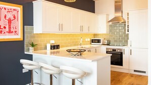 A modern kitchen with white cabinets, a central island with bar stools, a microwave, and a stainless steel range hood. - Stunning 2 Bed Apartment on the beach by Urban Roost (Worthing)