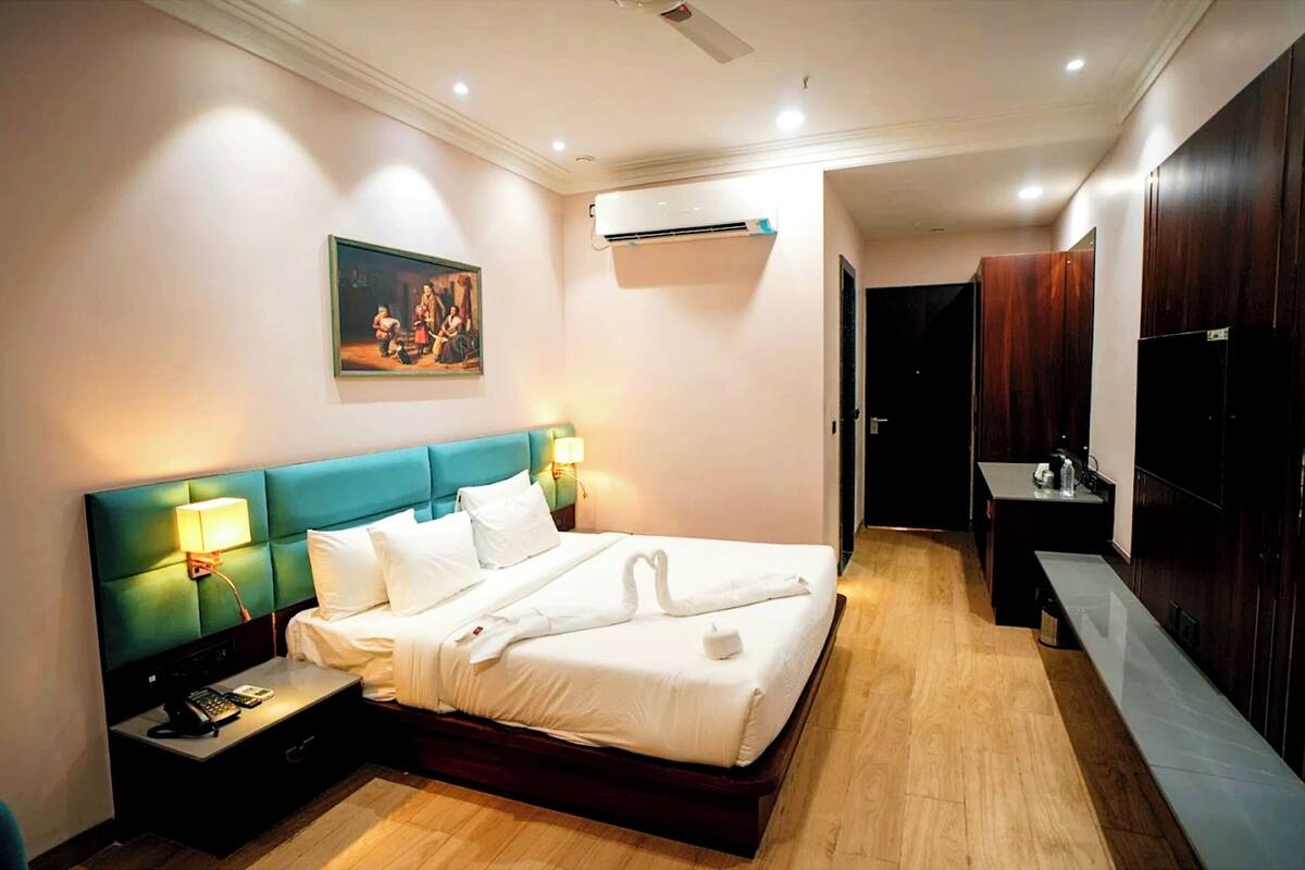 A hotel room with a bed, bedside tables, a painting, an air conditioning unit, and a wooden floor.