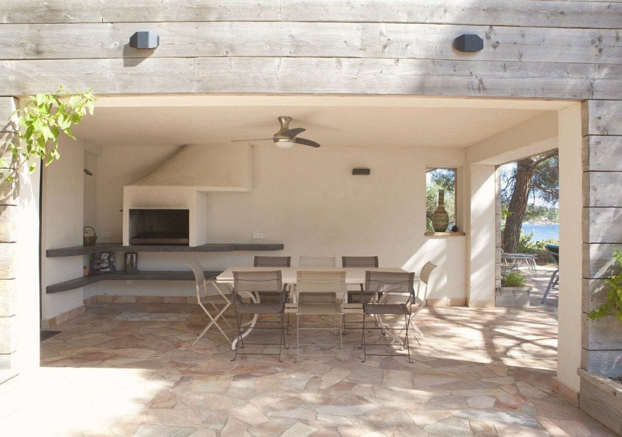 An outdoor patio with a stone floor, a table with chairs, a ceiling fan, and a view of the sea.