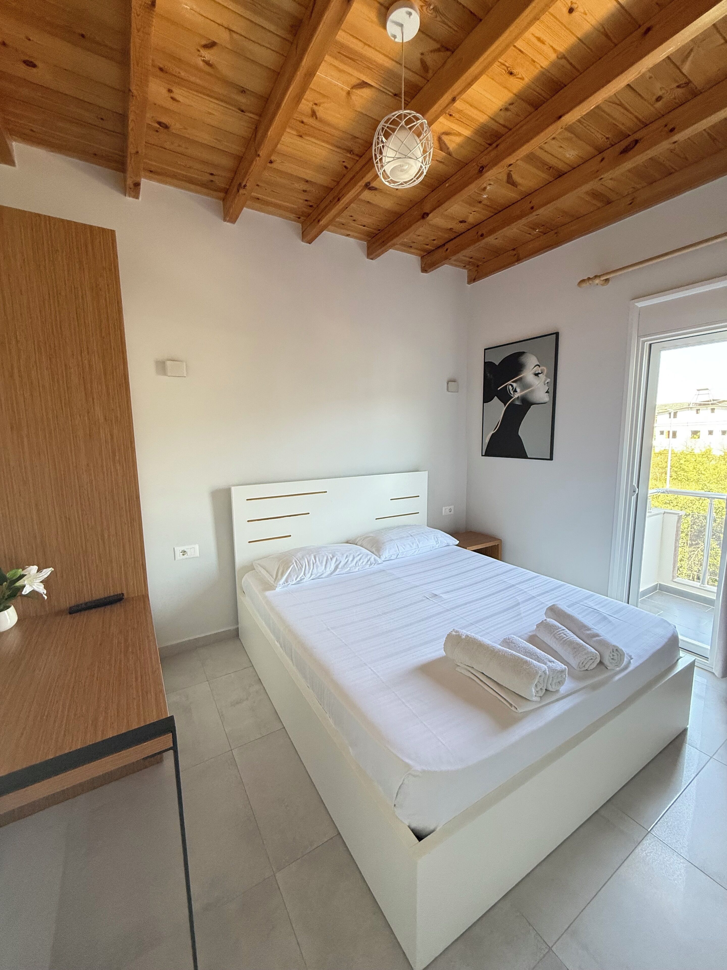 Quarto com teto de madeira, cama com roupa de cama branca, mesa de cabeceira, planta em vaso e janela com vista.