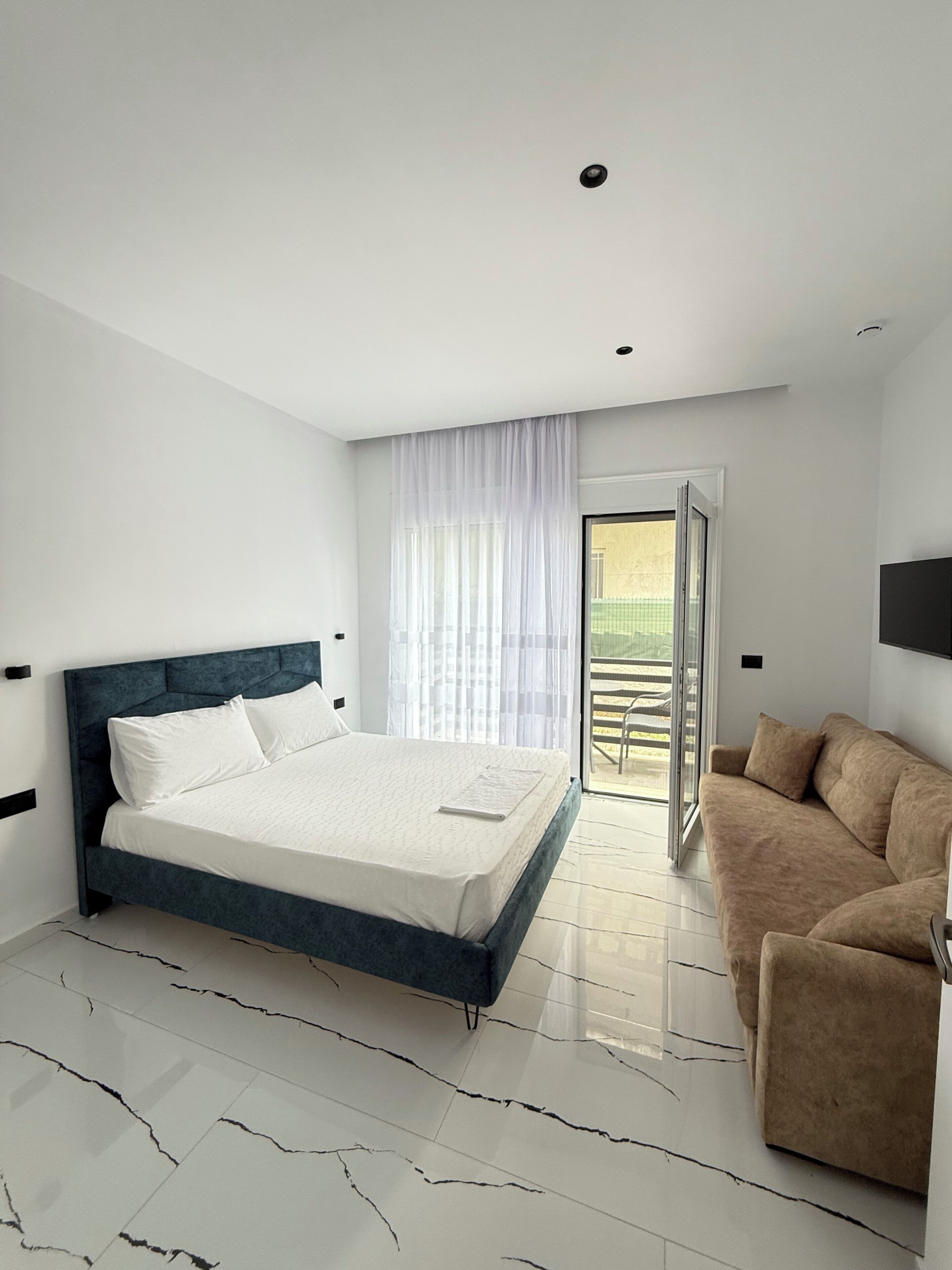 A modern bedroom with a large bed, a sofa, and a sliding glass door leading to a balcony.