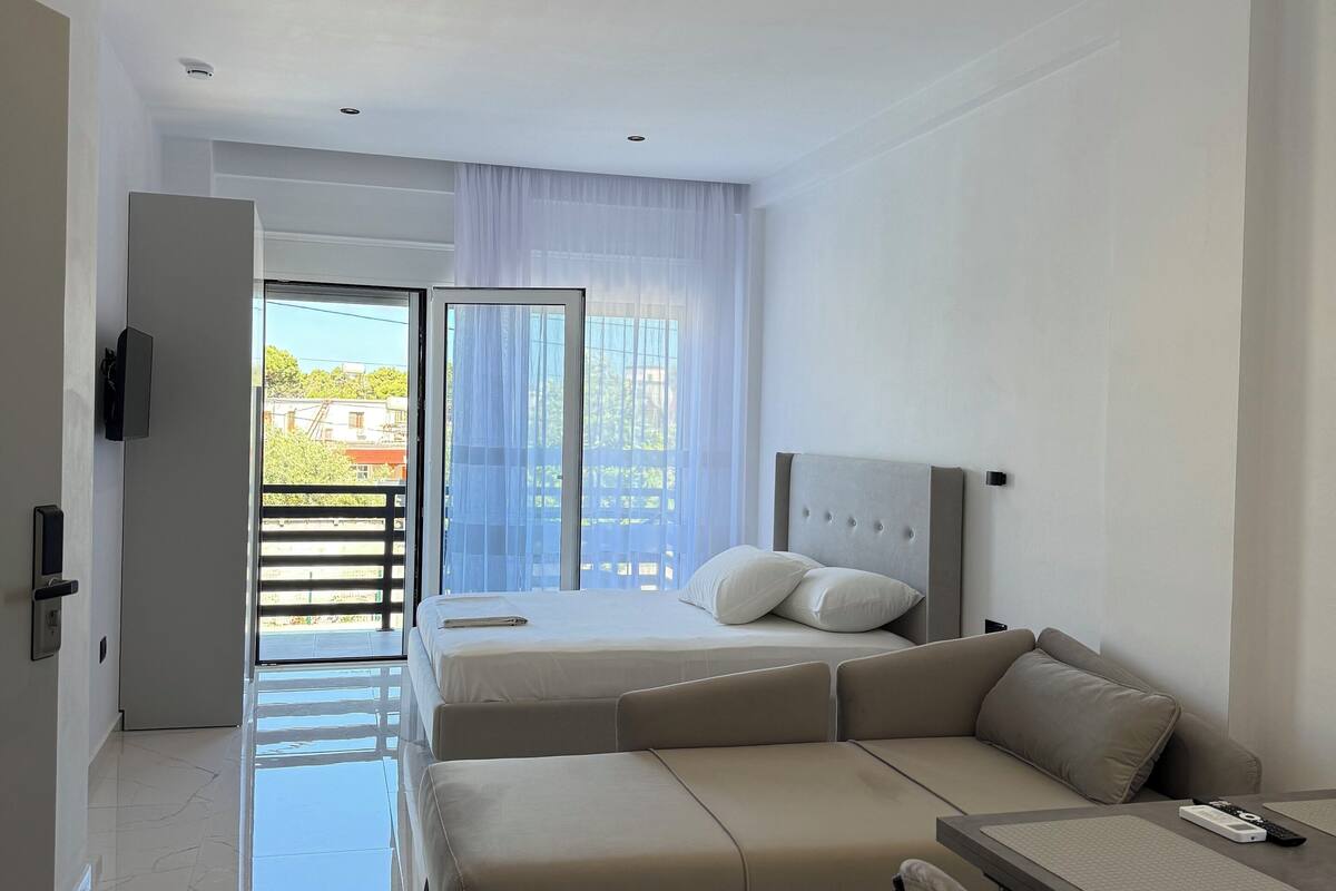 A modern hotel room with a large bed, a sofa, a dining table, and a balcony with a view of buildings.