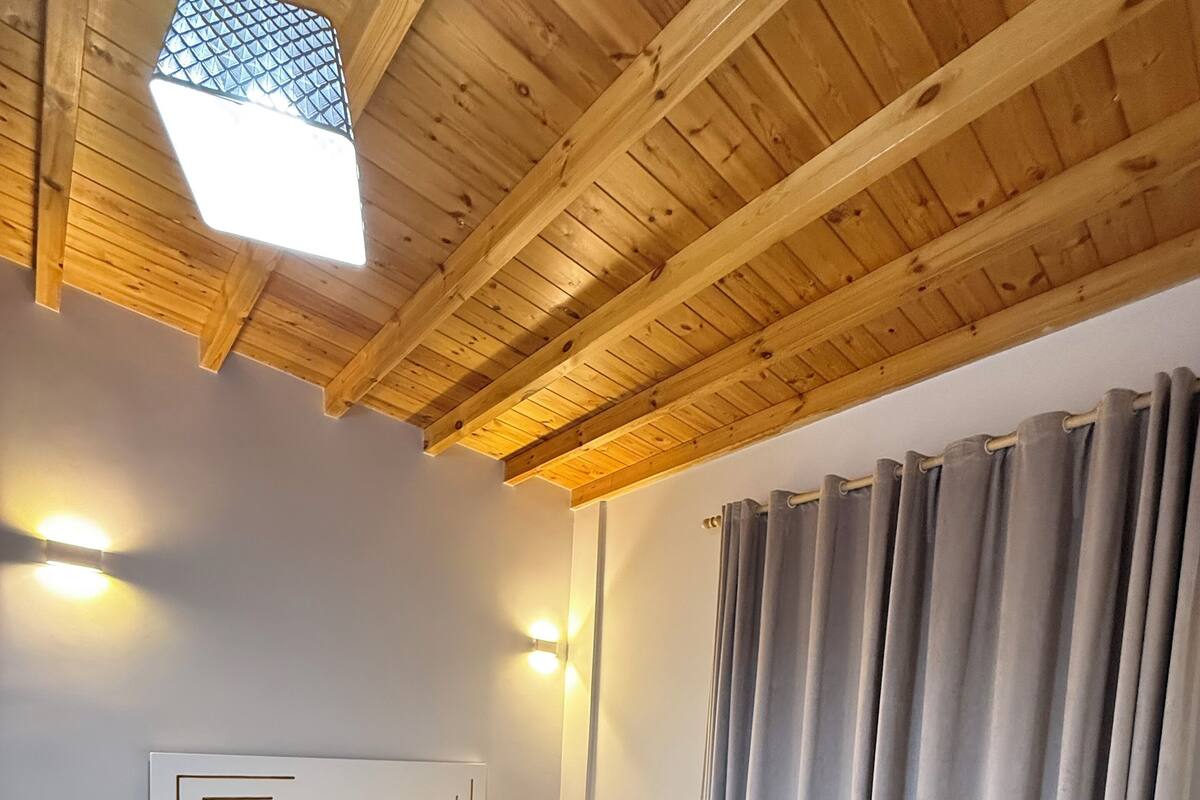 A bedroom with a wooden ceiling, a bed with white bedding, a bedside table, and a window with curtains.