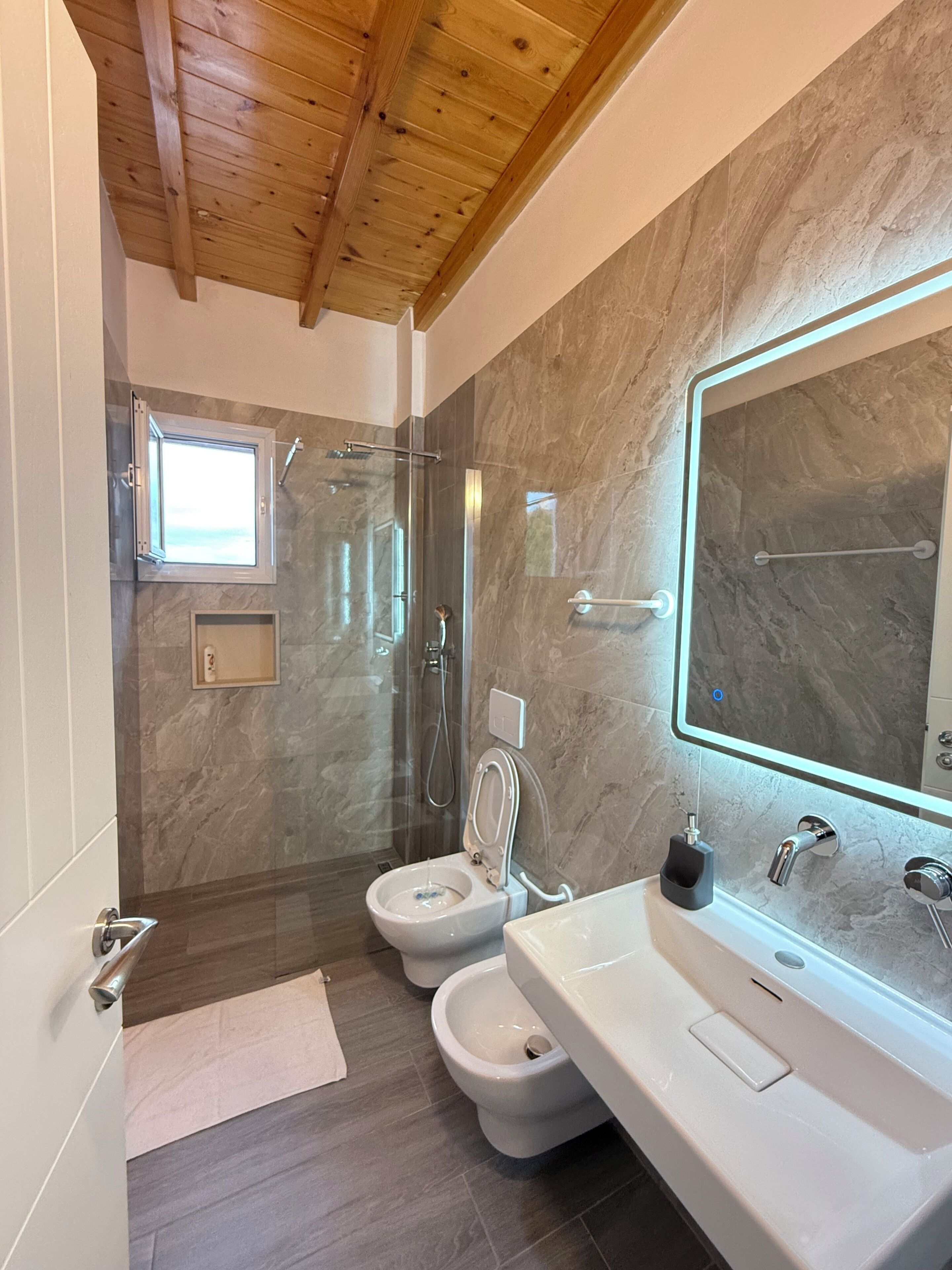 Un baño moderno con ducha de vidrio, dos inodoros, un espejo grande y un lavamanos.