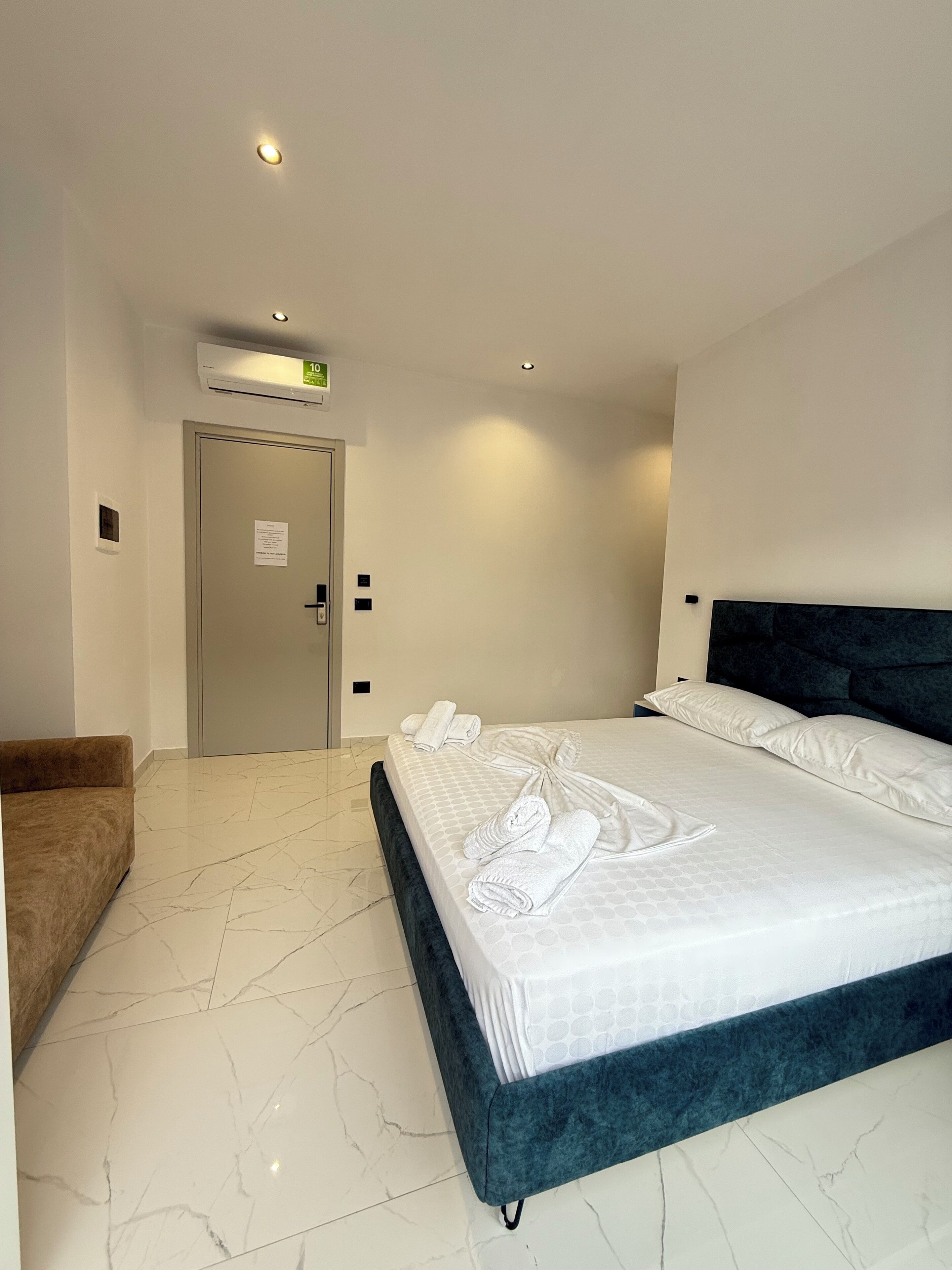 A hotel room with a large bed, a brown armchair, and a white air conditioning unit on the wall.