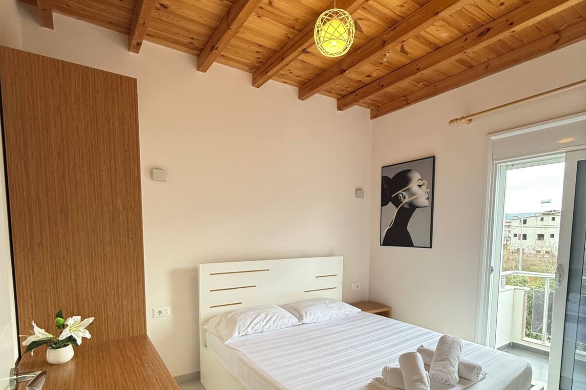A bedroom with a wooden ceiling, a bed with white bedding, a bedside table with a remote control, and a window with a view.