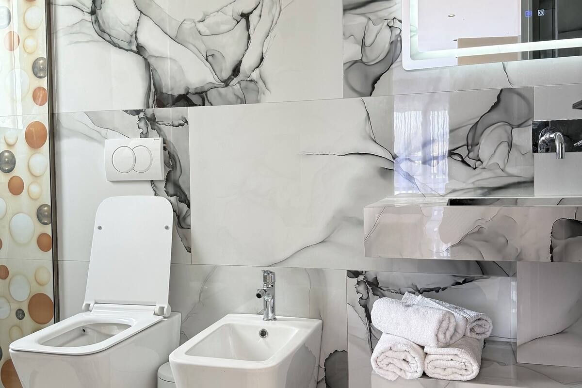 A bathroom with a toilet, bidet, and sink, all in white. There is a mirror above the sink and a towel rack with towels.