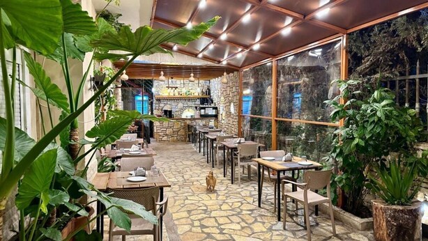 Een restaurant met stenen vloer, houten tafels en stoelen, en grote ramen met uitzicht op een stenen muur.