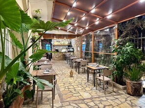 A restaurant with stone flooring, wooden tables, and chairs, and large windows overlooking a stone wall. - Maison Apartments (Vlore)