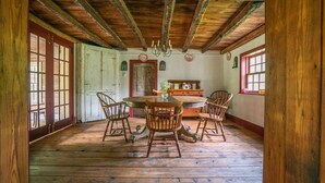 Een rustieke eetruimte met een houten tafel en stoelen, een kroonluchter, en uitzicht op het exterieur door grote ramen.
