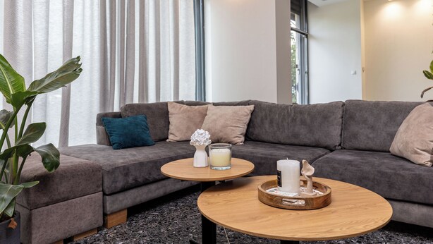 A modern living room with a grey sofa, a round wooden coffee table, and a potted plant. - Nobia 3 (Athens)