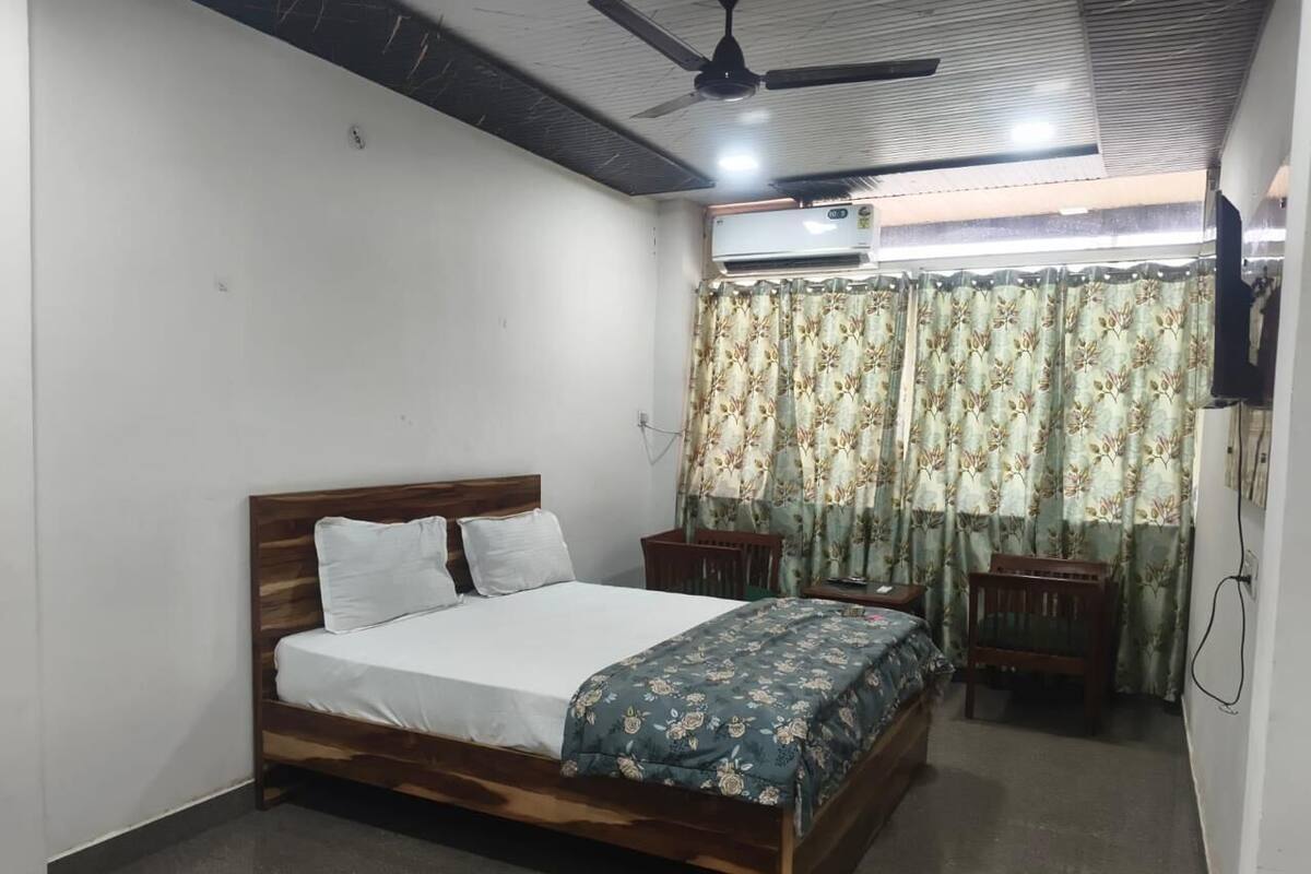 A bedroom with a wooden bed, a ceiling fan, an air conditioning unit, a small wooden table, and a curtain with a floral pattern.