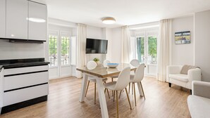 A modern kitchen with white cabinets, a black countertop, and a built-in oven. There is a dining area with a wooden table and white chairs. A flat-screen TV is mounted on the wall above the table. - Stay U-nique Apartment Plaza de la Merced (Málaga)