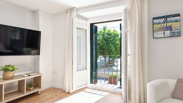 A modern living room with a flat-screen TV, a wooden console table with plants, a white sofa, and a sliding glass door leading to a balcony with greenery. - Stay U-nique Apartment Plaza de la Merced (Málaga)