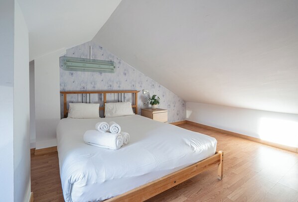 A bedroom with a wooden bed, white bedding, a nightstand, a lamp, and a potted plant.