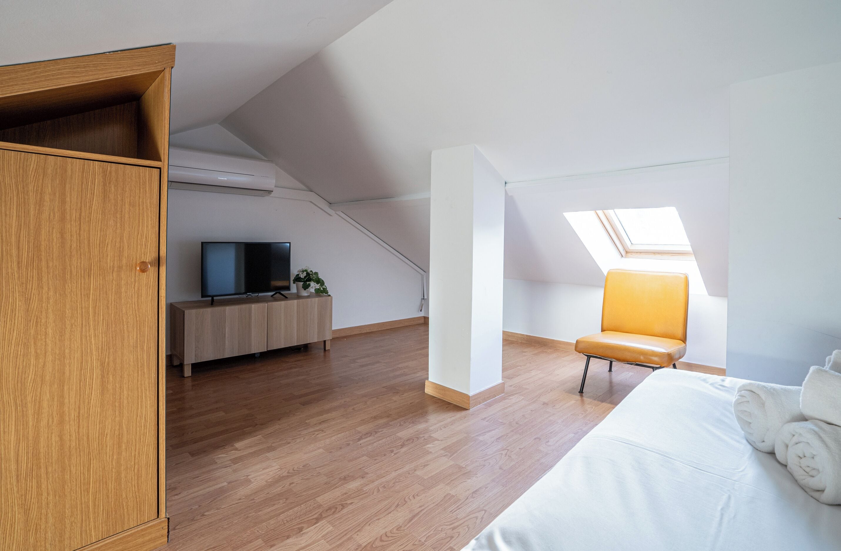 A modern, well-lit room with a wooden wardrobe, a bed with white linens, a television on a wooden stand, and a skylight.