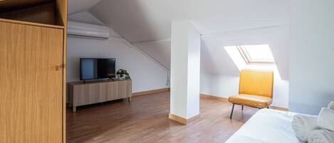 A modern, well-lit room with a wooden wardrobe, a bed with white linens, a television on a wooden stand, and a skylight.