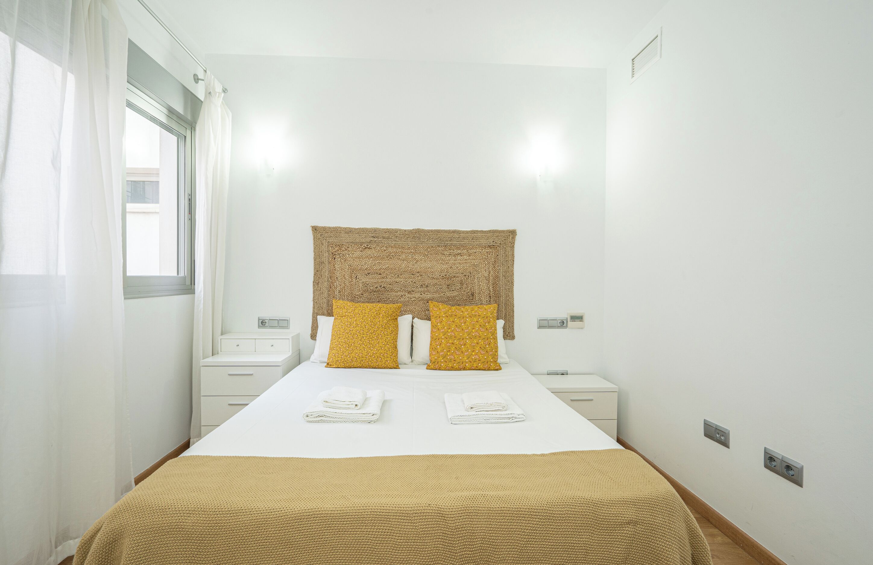 A hotel room with a bed, two bedside tables, a headboard, and a window with curtains.