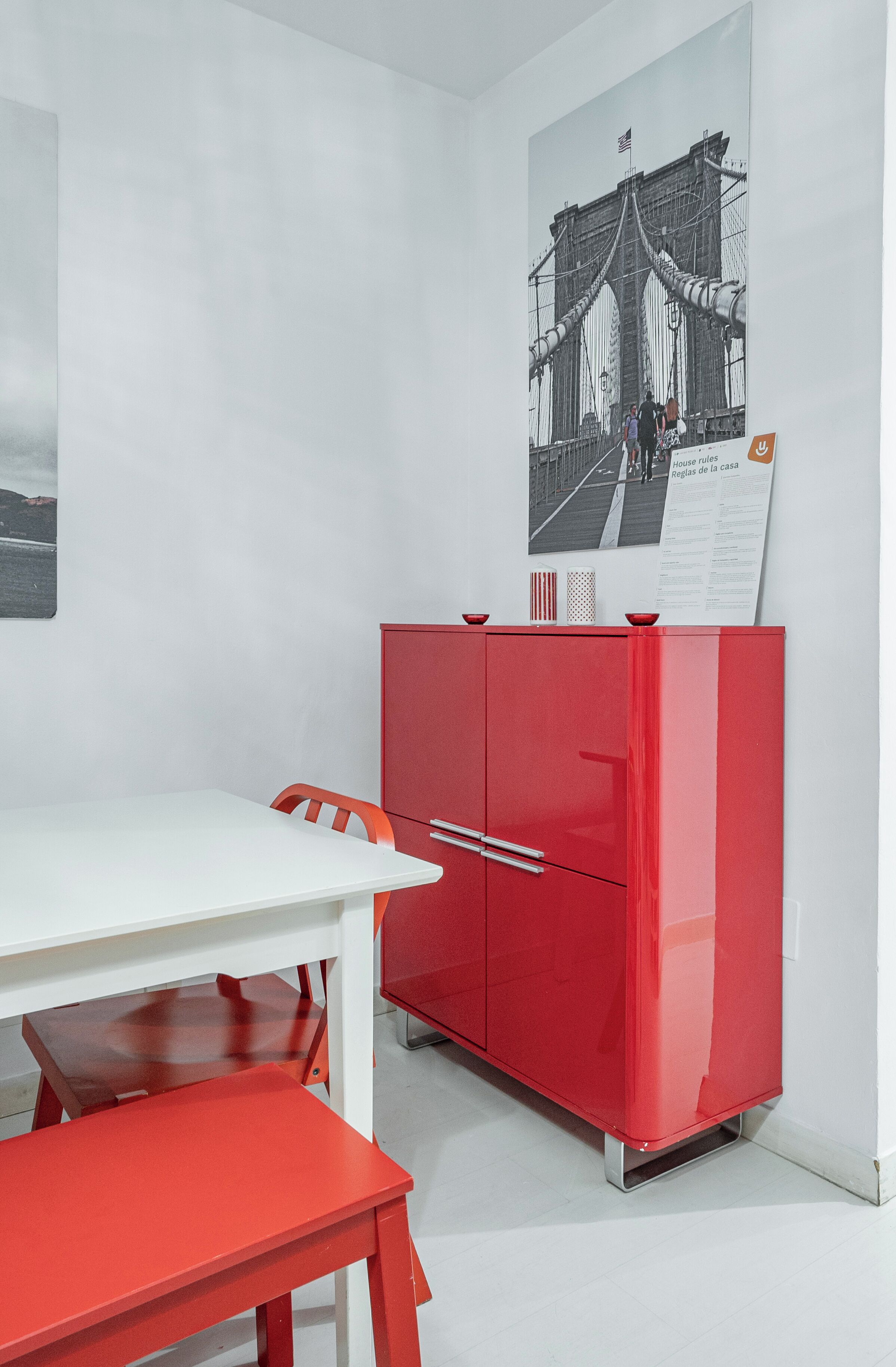A room with a white table and red chairs, a red cabinet, and a poster of a bridge.