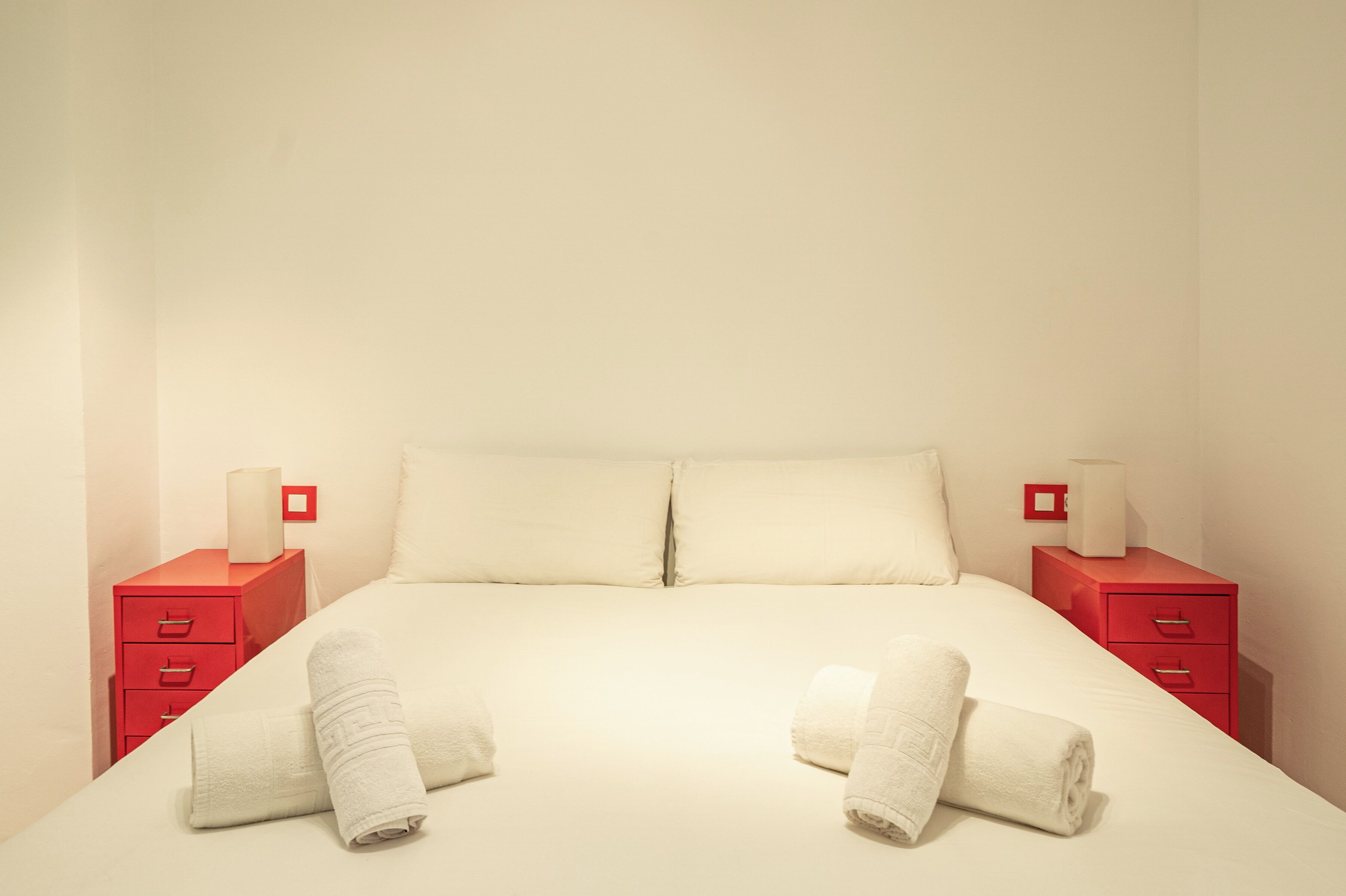 Un lit bien fait, avec deux oreillers, deux tables de chevet rouges surmontées de lampes blanches, et deux serviettes enroulées posées sur le lit.
