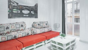 Un salon moderne avec un canapé blanc, des coussins rouges, une table basse blanche et un tableau représentant un paysage urbain accroché au mur.