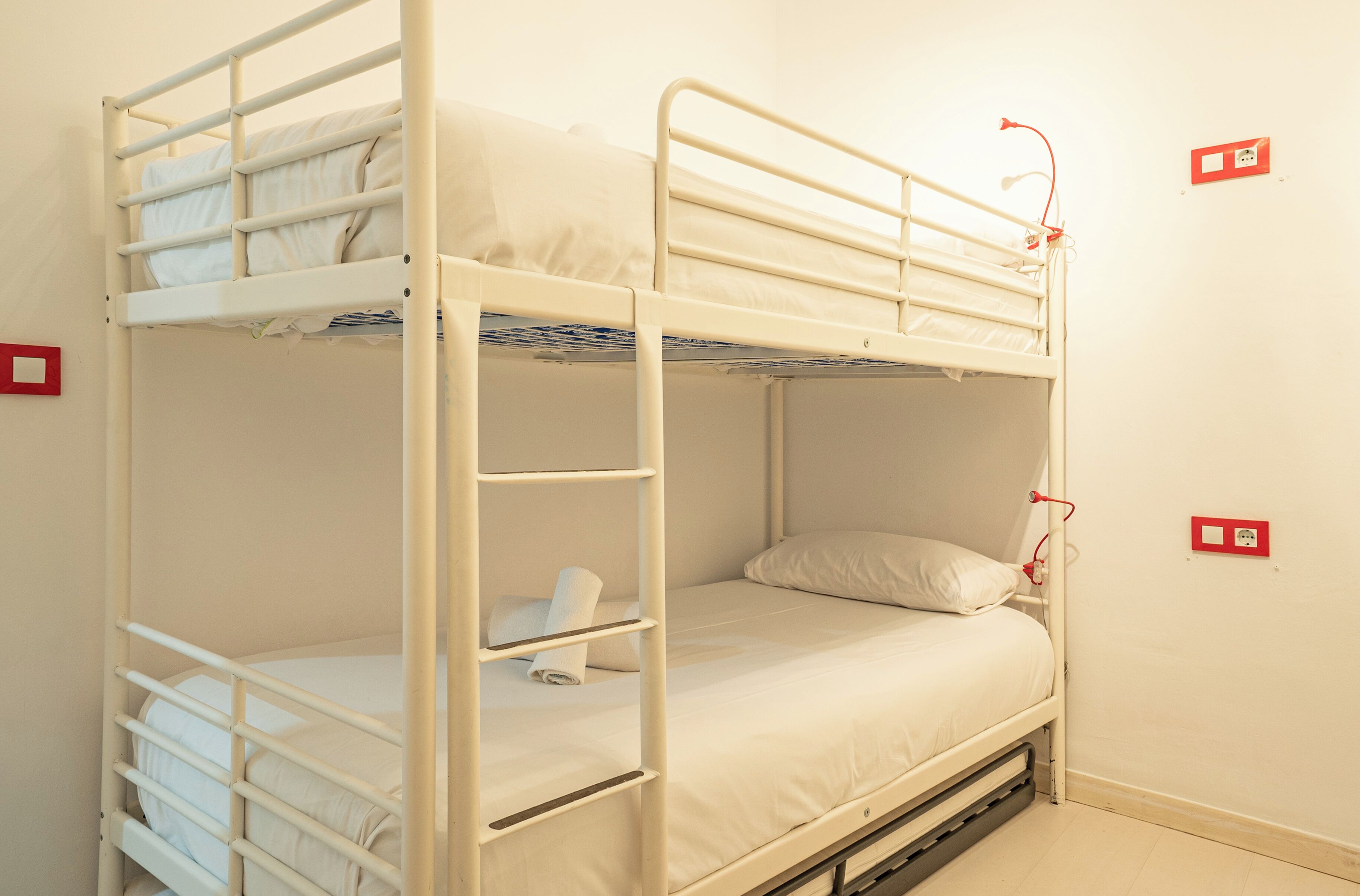 A bunk bed with a ladder, a single bed below, and a red emergency light on the wall.